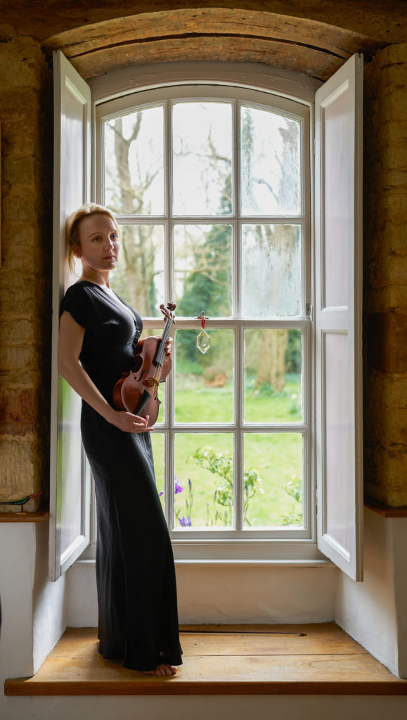 May Robertson in a black dress holding her violin with both hands by a window